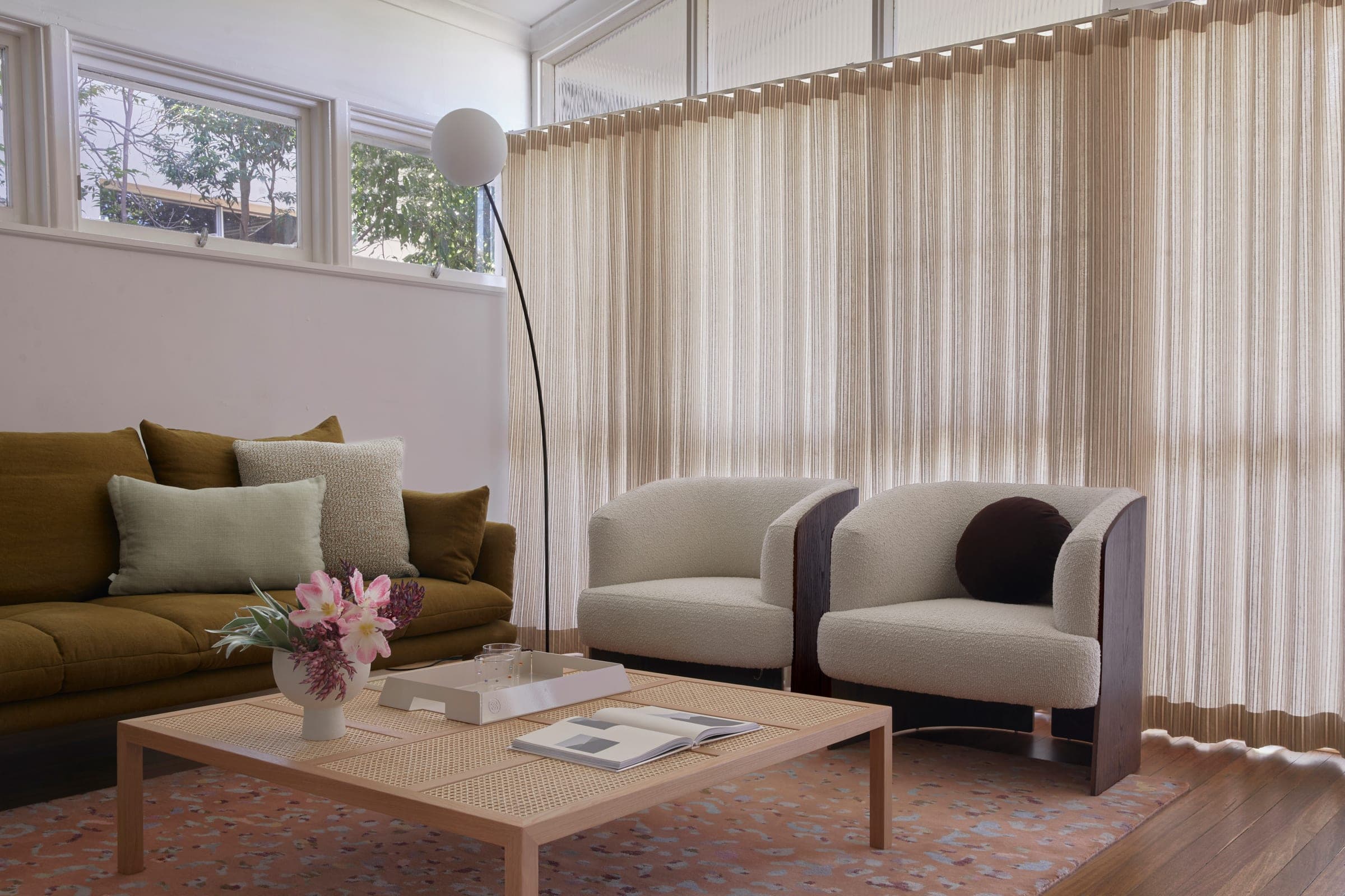 Living room with olive sofa, two boucle armchairs, cane coffee table and arc floor lamp, on a terrazzo-pattern rug.