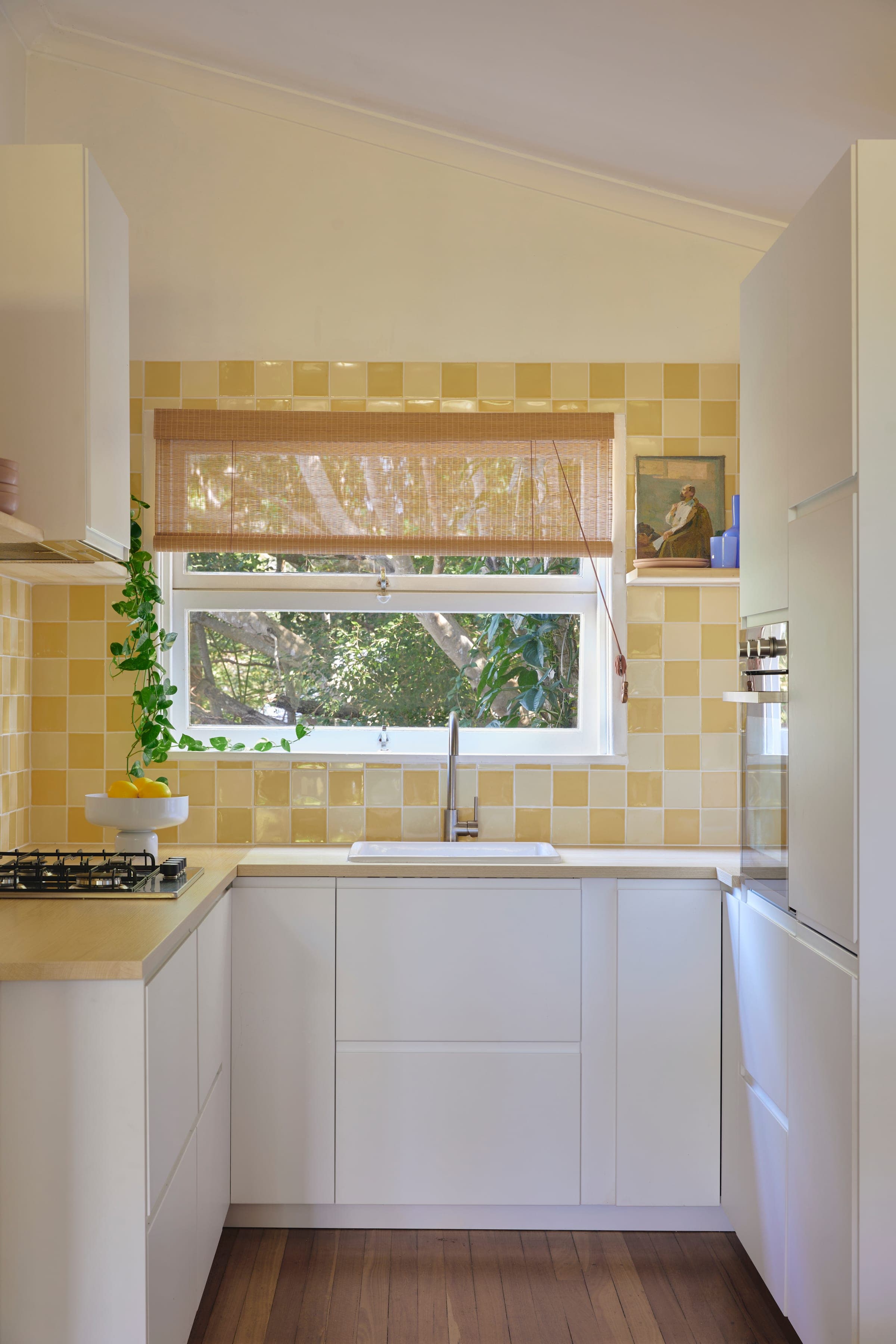 Yellow checkerboard tile splashback frames the kitchen window with a woven blind and view to a fig tree.