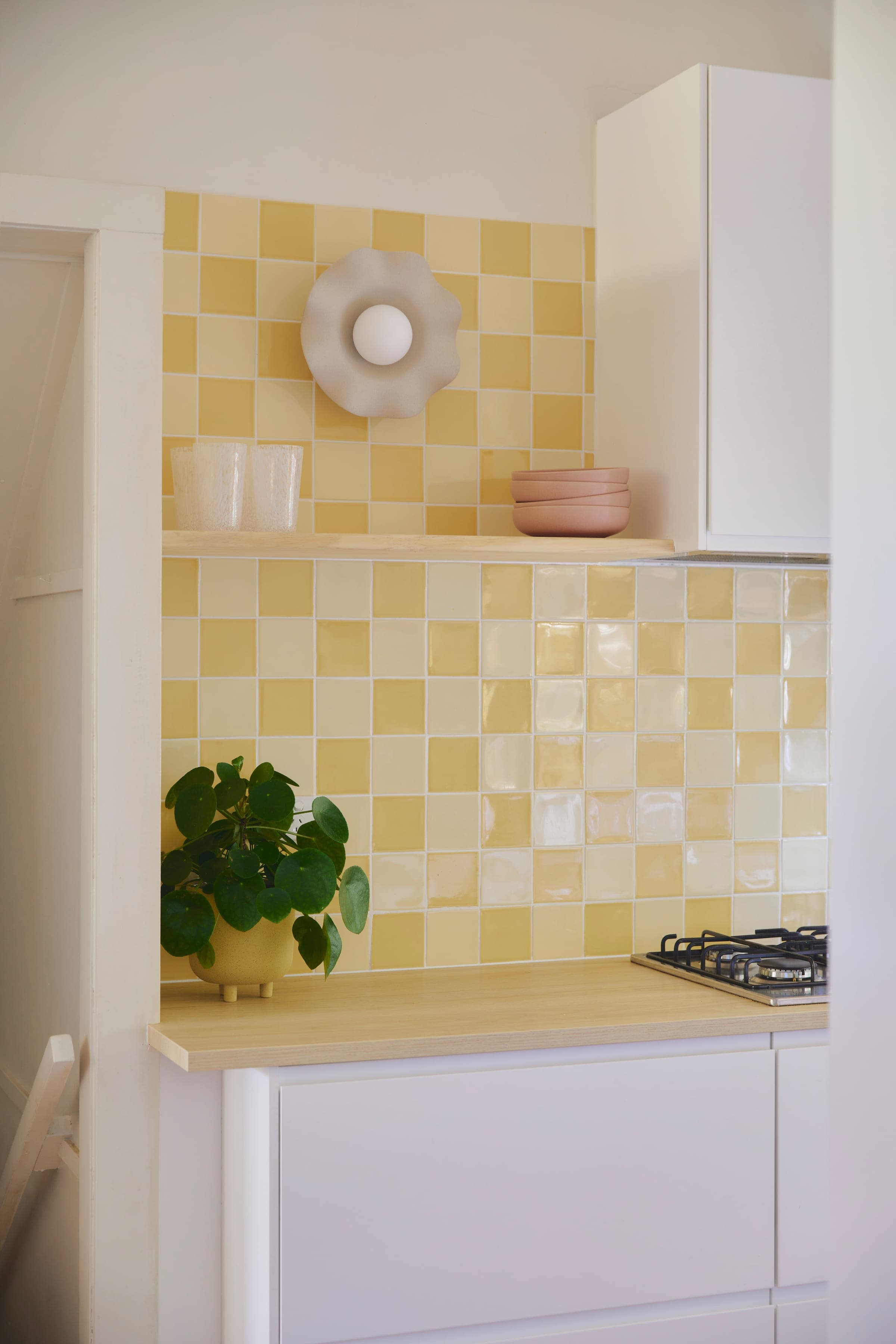 Open kitchen shelf with a sculptural ceramic vase, dirty-pink stacked bowls and the yellow checkerboard tile behind.