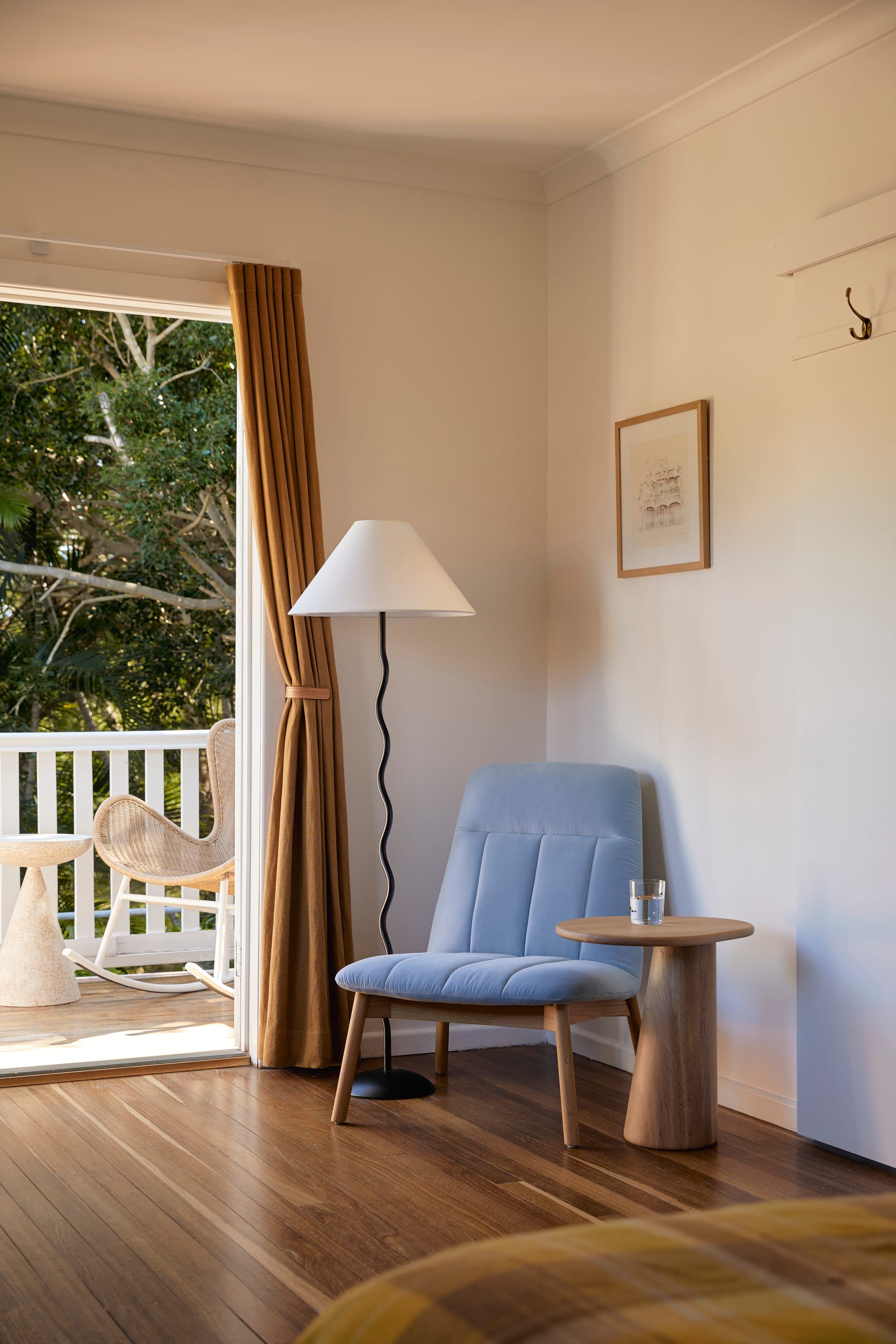 Bedroom reading corner with pale blue armchair, wooden side table and a glimpse through gold curtains to the balcony.