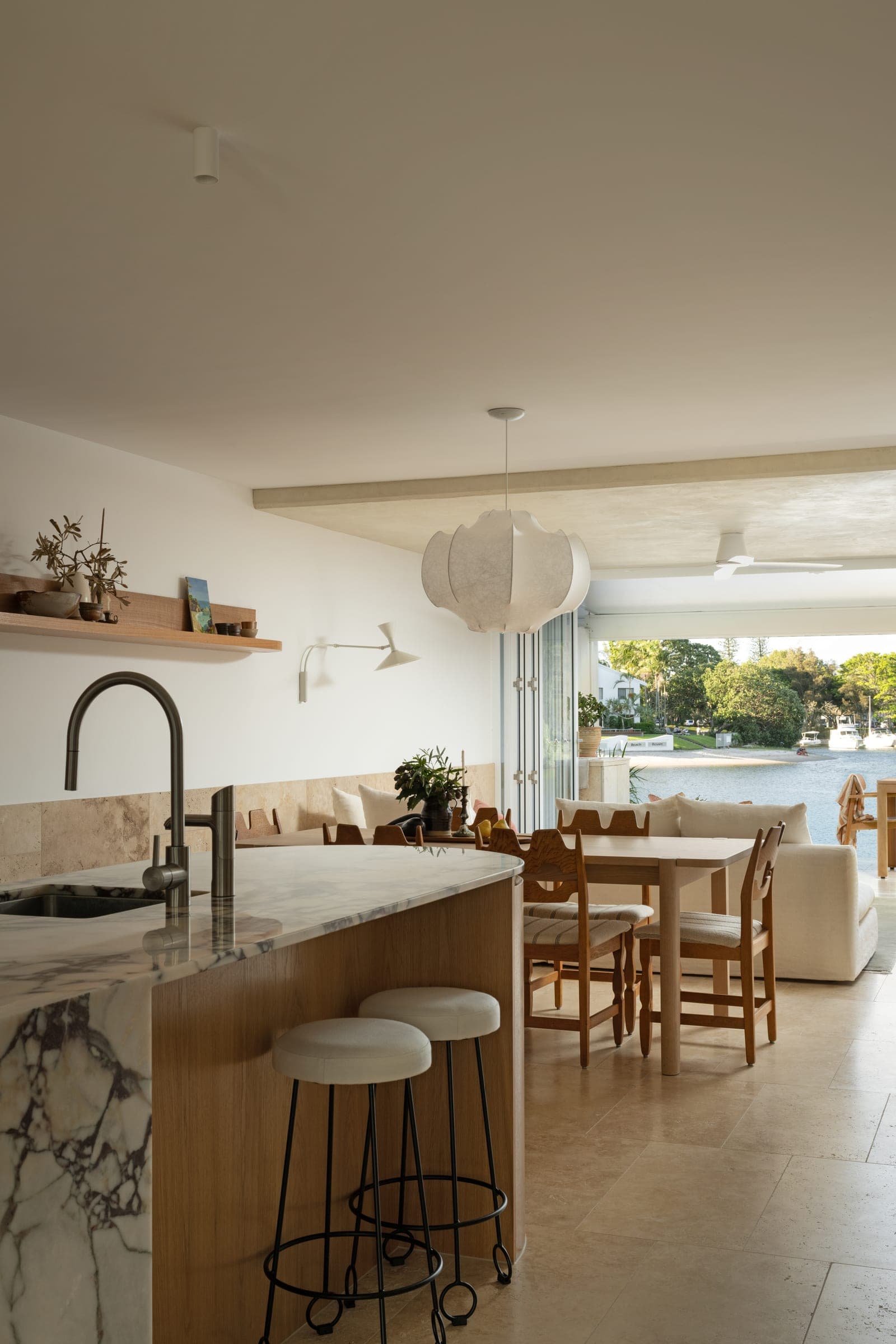 Kitchen looking toward the dining table and water, with a curved veined-marble island, oak joinery and a travertine dado wrapping the back wall.