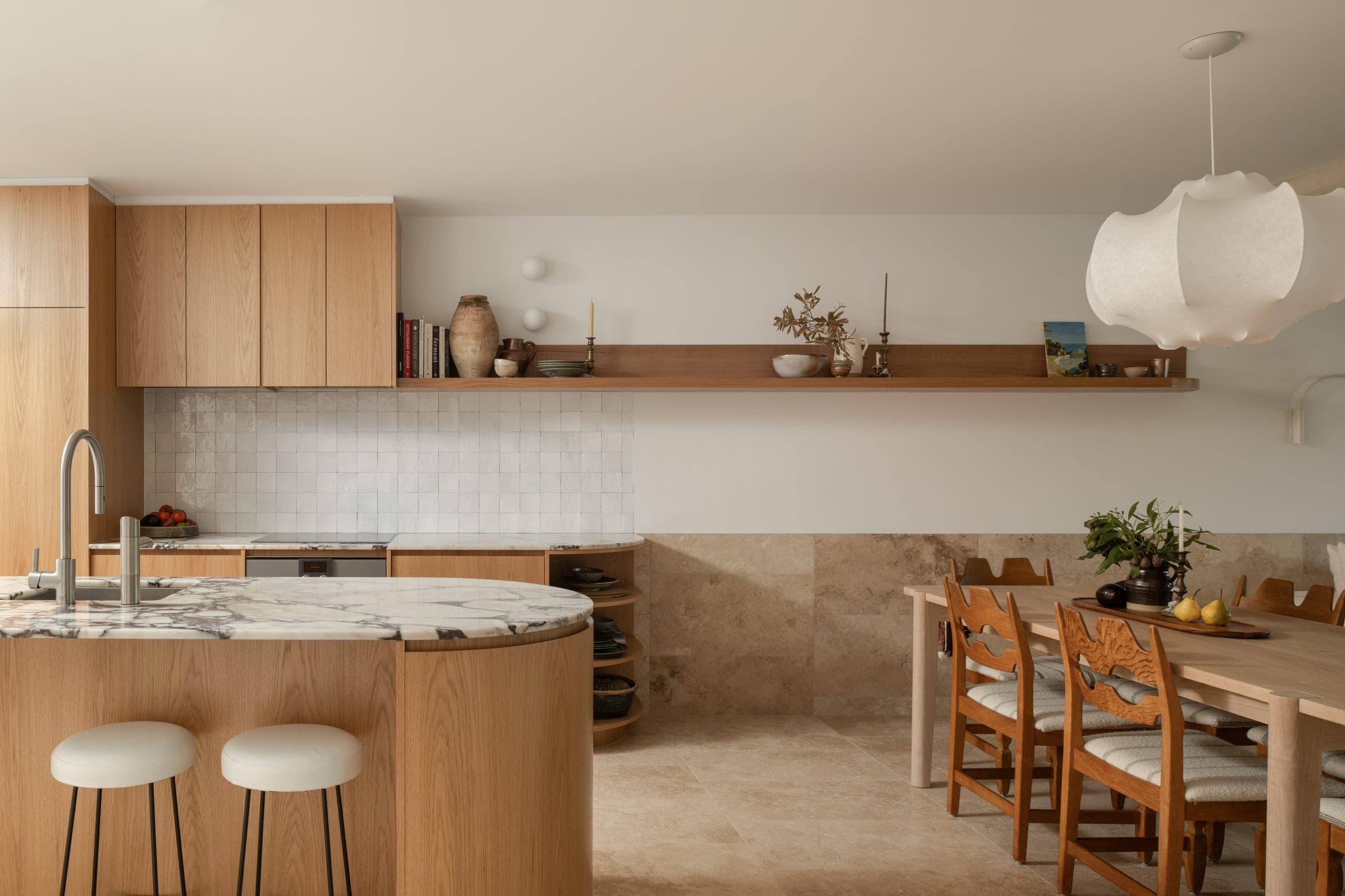 Kitchen and dining room with zellige splashback, oak upper cabinetry, travertine dado, and high-back oak dining chairs around a long timber table.