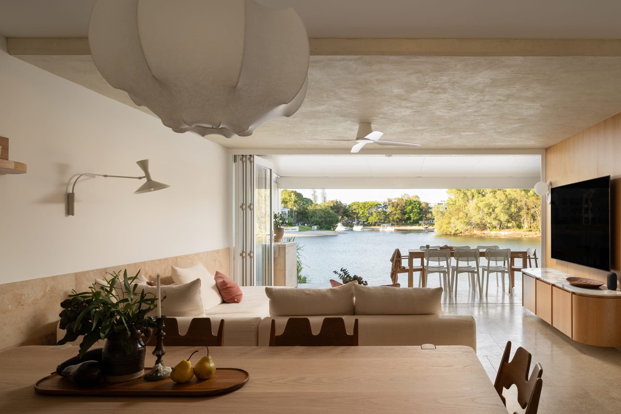 Living room with a large paper lantern pendant, linen sofa and oak dining set, looking through bifolds to the river and trees beyond.