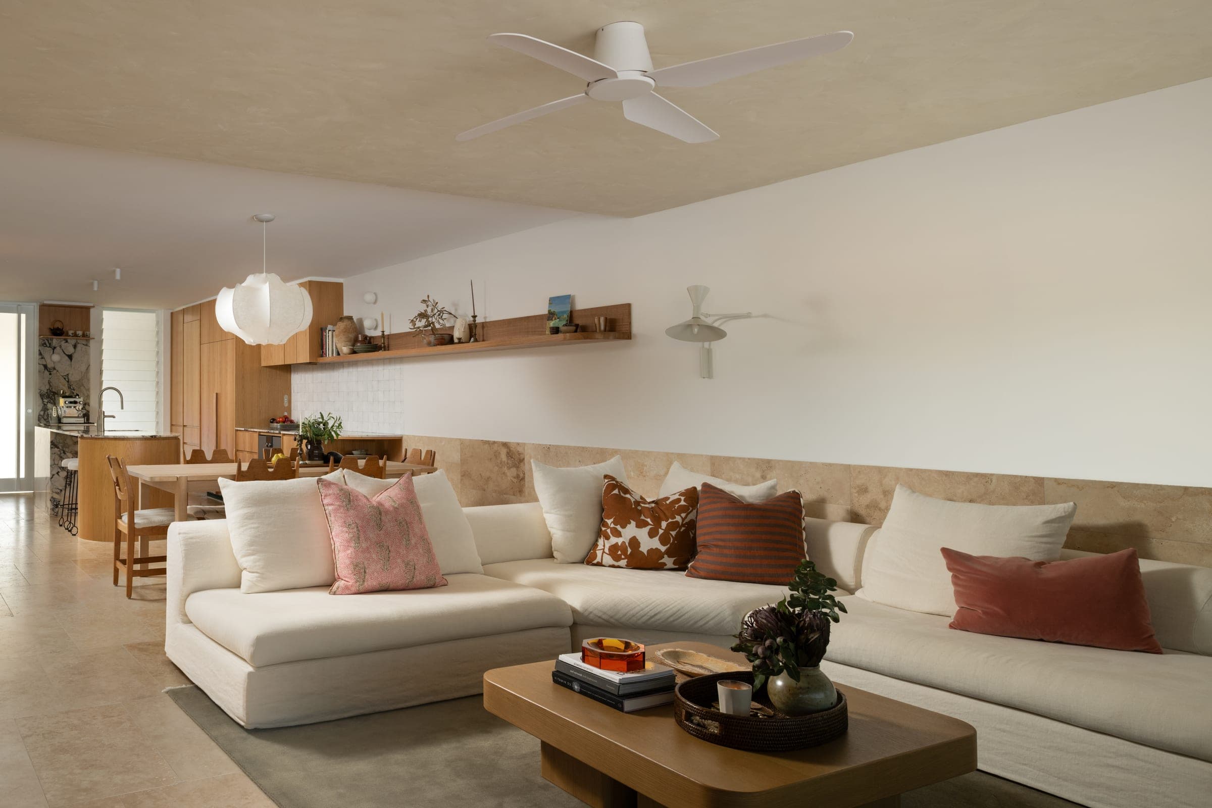 L-shaped linen sectional banked against a travertine dado, layered in rust, pink and striped cushions, with the oak kitchen and dining table visible beyond.