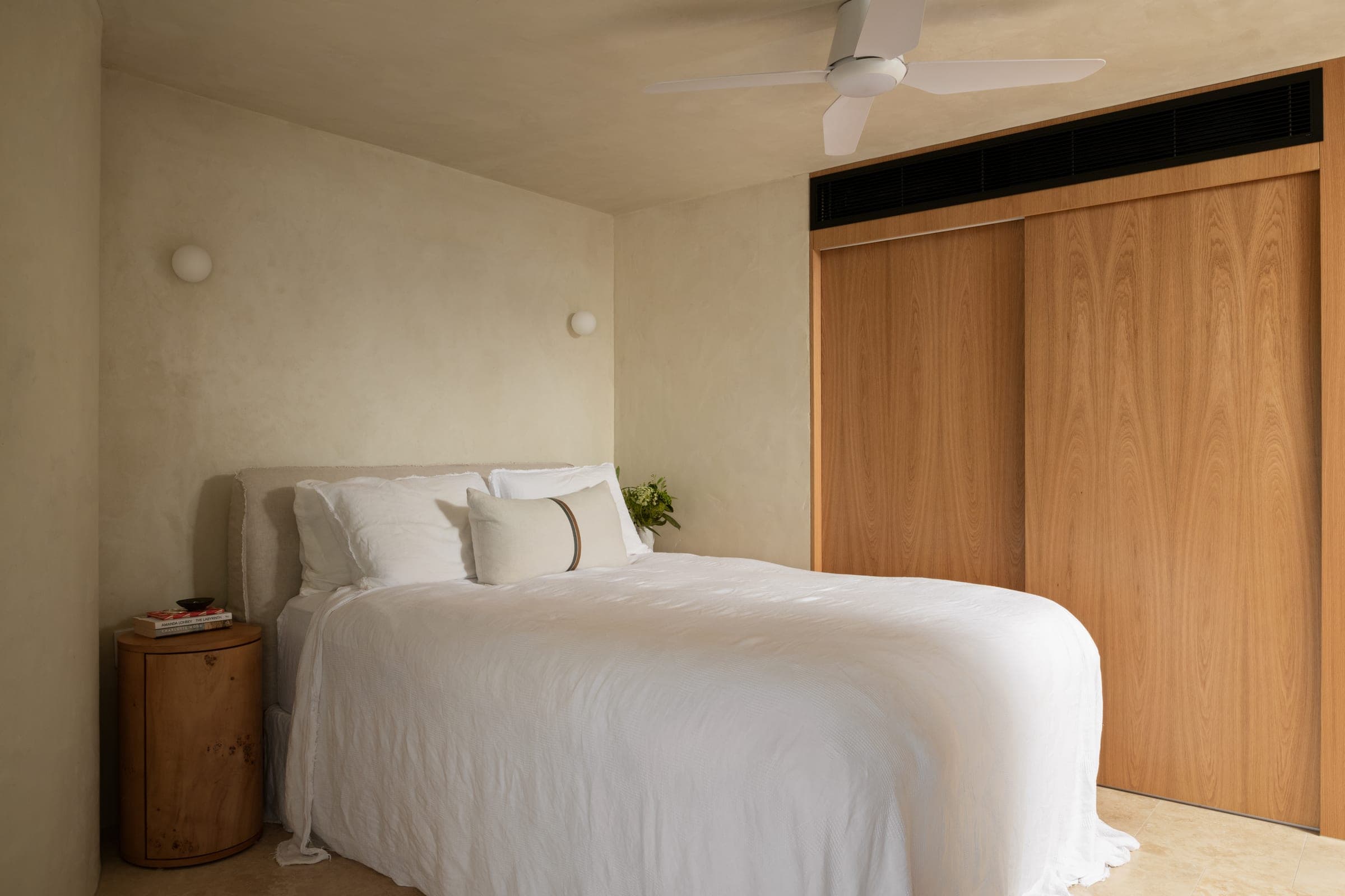 Second bedroom with a low linen-upholstered bed, full-height oak sliding wardrobe and a turned burl-timber drum side table on travertine floor.