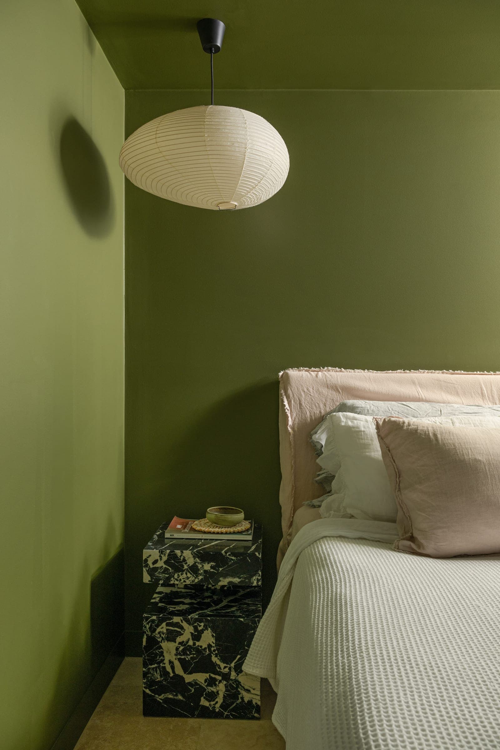 Bedroom in deep olive green, with a linen-upholstered bed, paper lantern pendant overhead and stacked dark-marble cube side tables.