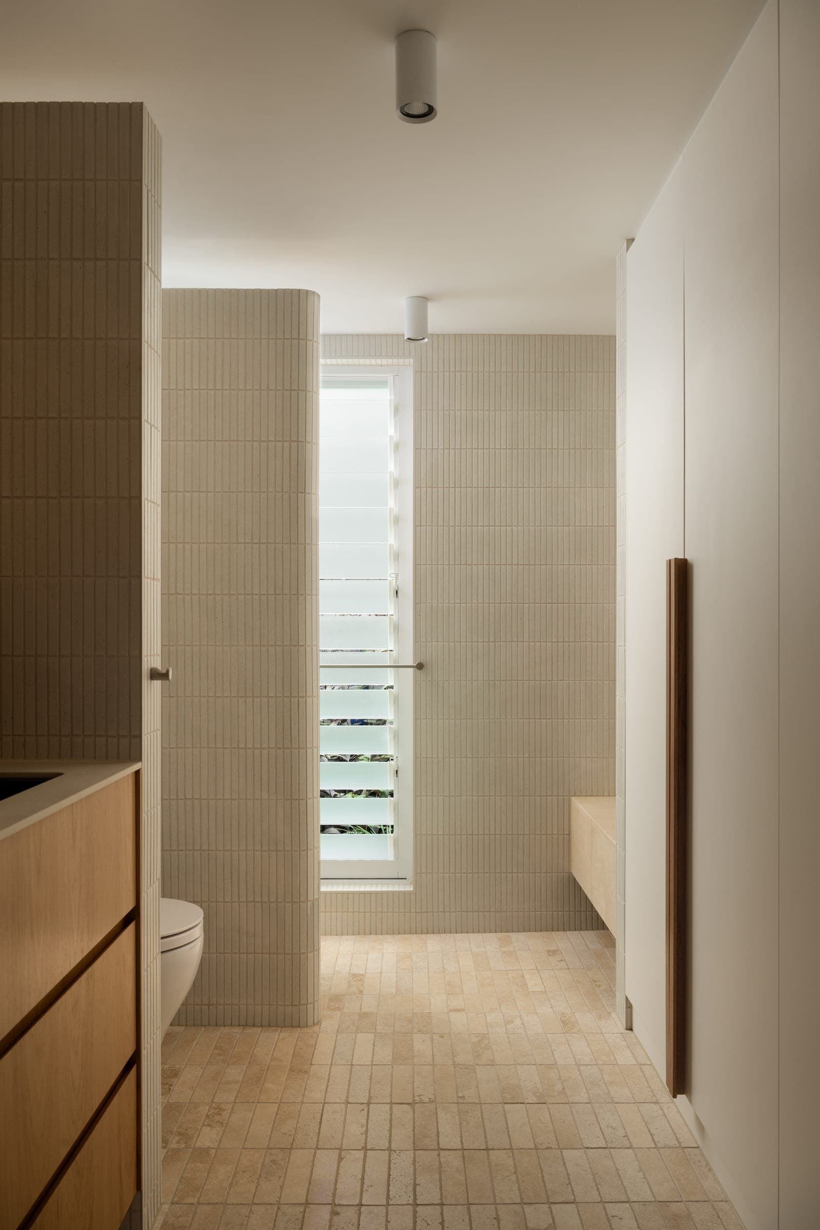 Ensuite corridor with cream brick-format tile, travertine floor and a slim window framing greenery beyond.