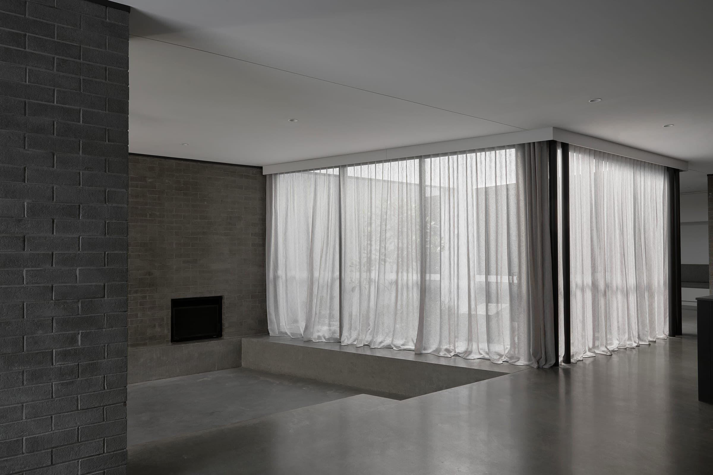 Living room with grey brick fireplace, polished concrete floors and full-height white sheer curtains.