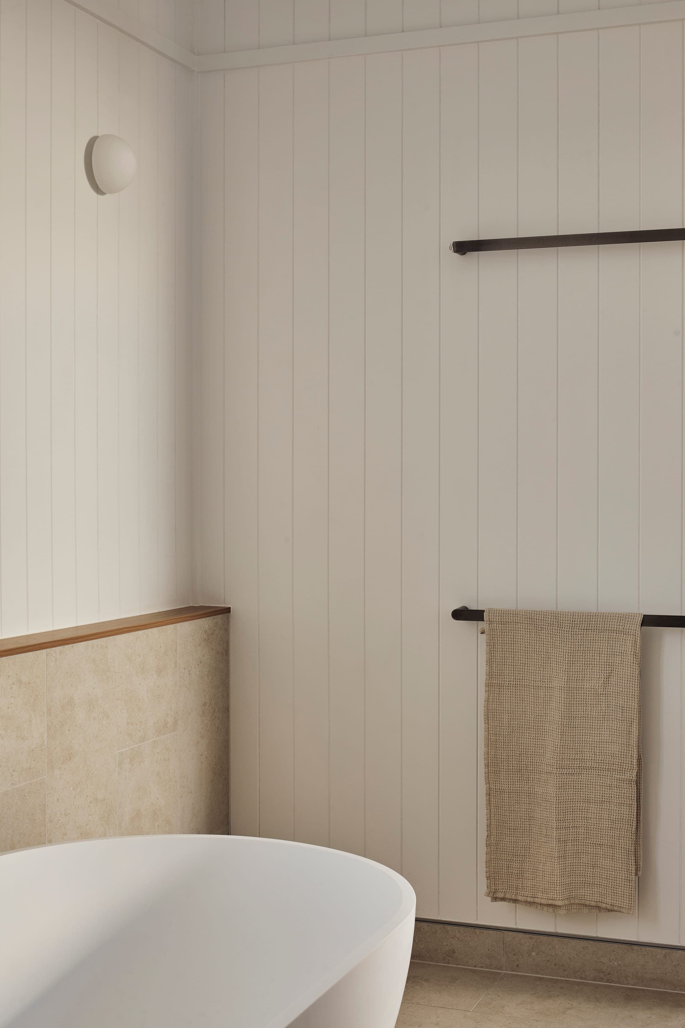 Bathroom corner with vertical shiplap, beige stone tile dado and leather towel rails next to a freestanding tub.