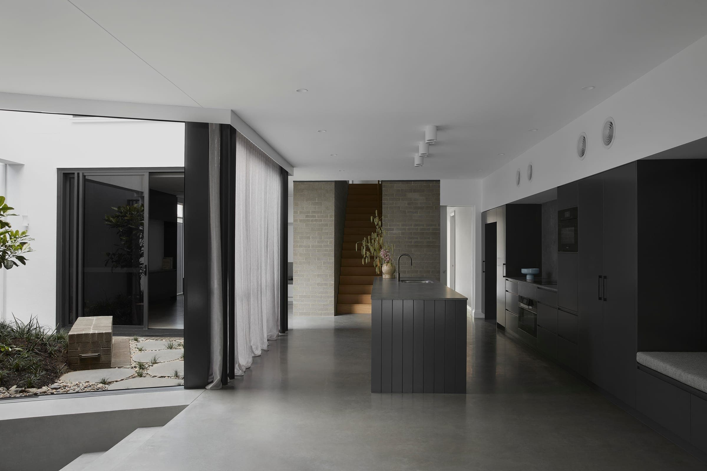 Open-plan kitchen with black island and dark joinery, looking through sliding doors to a paved courtyard with a rubber plant.