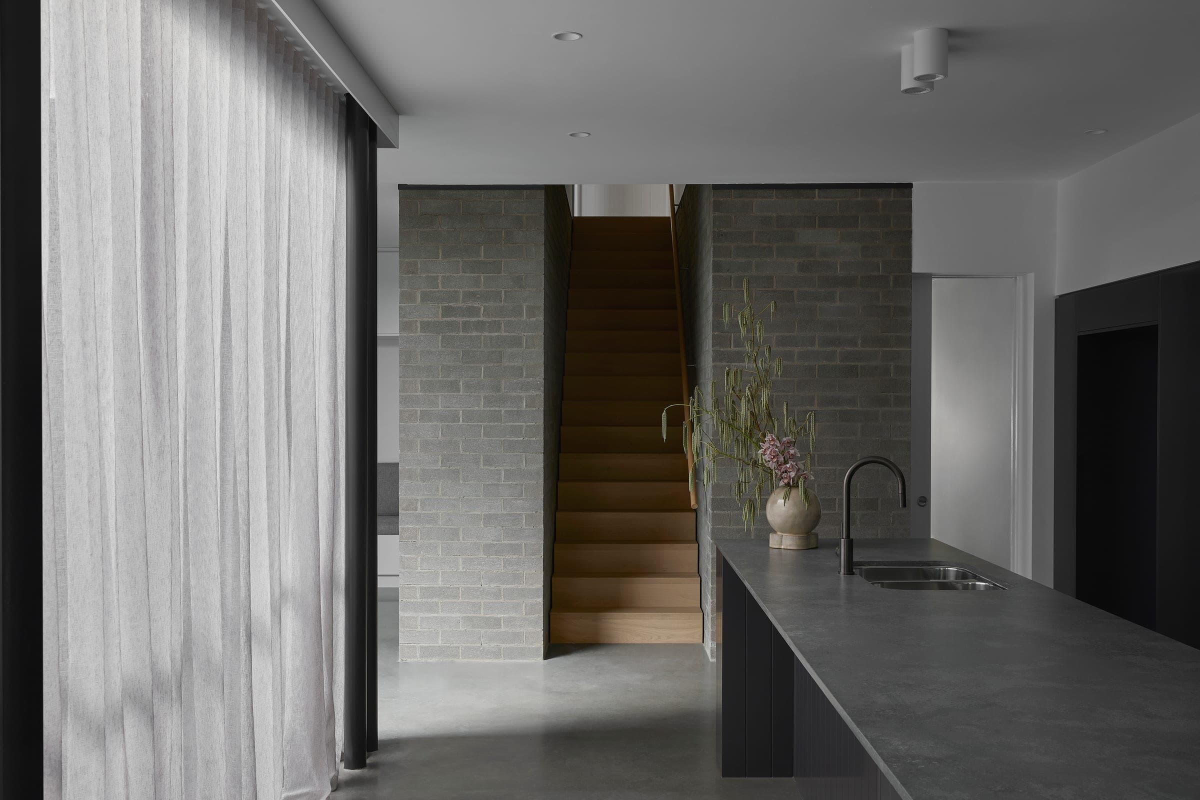 Kitchen sequence with full-height sheer curtains, a beige brick stair pillar and the dark island bench to the right.